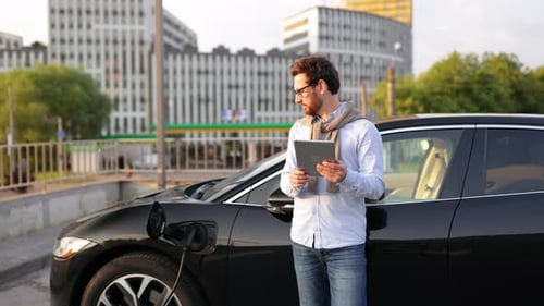 Man Uses Tablet near Electric Car Charging