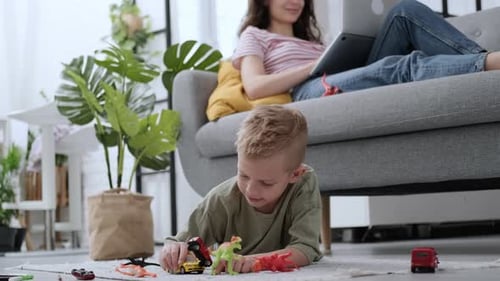 Little Boy Playing on Floor Dinosaur and Cars Toys While Mother Working Remotely From Home on Laptop
