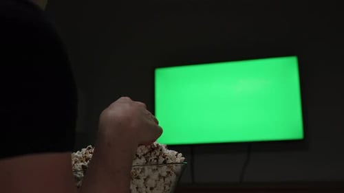 In a dark room in front of a green screen television, a woman eats popcorn.