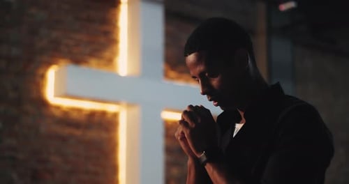 Man Praying by Luminous Cross in Dark Room