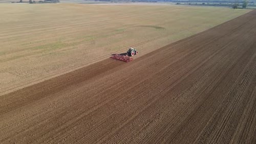 An aerial footage of a tractor working on a large agricultural field, plowing the soil in straight,