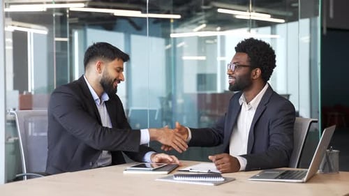 Two businessmen in formal suits shaking hands after successful negotiation while sitting at office
