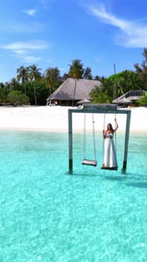 Woman Swings Above the Crystal Clear Sea on a Wooden Swing Facing the Beach and Lush Tropical