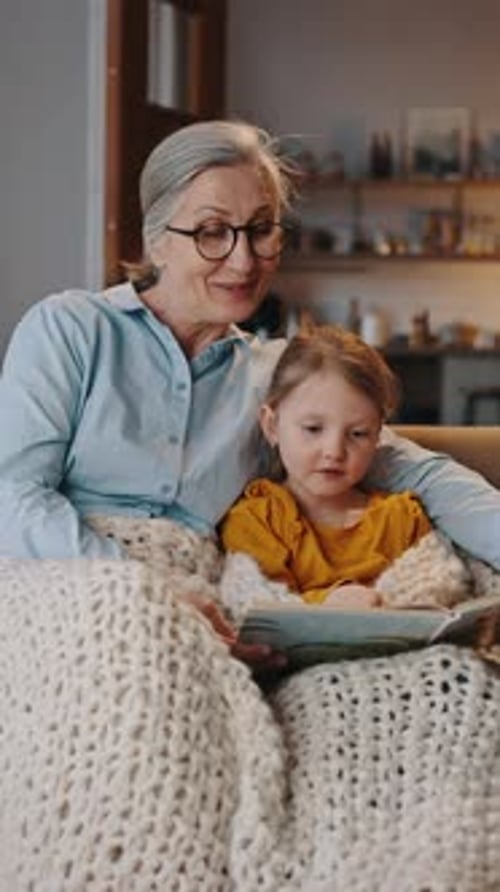 Grandmother Reading Book to Granddaughter Indoors Under Blanket