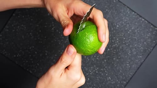 Cutting fresh lime by hand. Hands cutting fresh green lime with knife on dark cutting board.