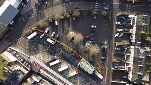 A slow, forwards flying top down shot, of a small city bus depot over a road and some buildings.