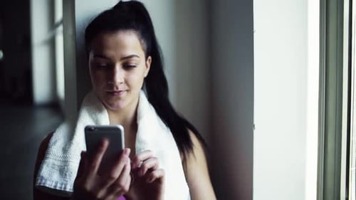 Young woman in gym sits resting and texting on her smartphone after workout