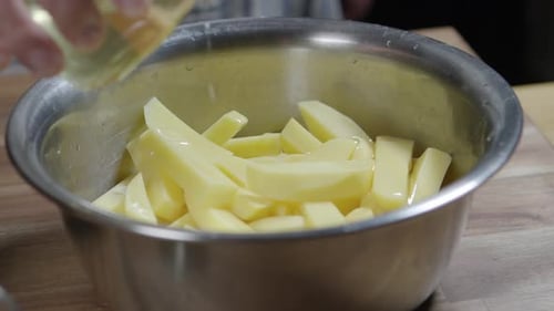 A chef pours olive oil over raw potato wedges in a metal bowl.