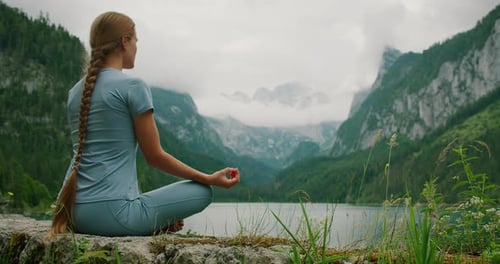 Woman meditates by a peaceful lake, surrounded by majestic mountains and clouds, immersed in deep re