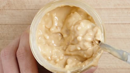 Stirring peanut butter in a jar with a spoon, top view on a wooden table. Close-up.