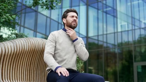 A sick businessman suffering from a sore throat while sitting on a bench on the street near business