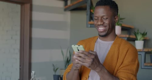Cheerful African American Man Using Smartphone Messaging in Social Media Holding Gadget Indoors in