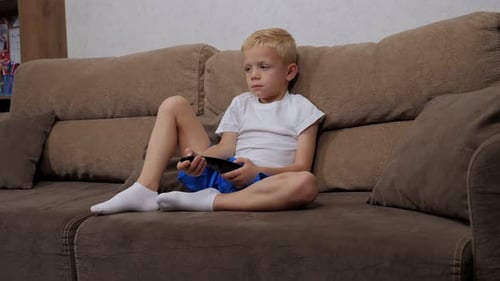 Der kleine Junge sitzt auf der Couch und wechselt mit der Fernbedienung zwischen den Fernsehkanälen