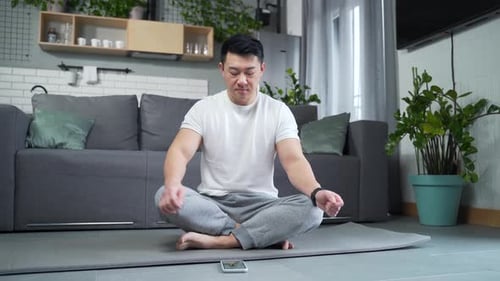 Asian man practicing yoga sitting on mat in lotus position at home in living room. meditates