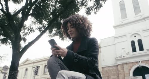 A curly woman in a suit sits in the park while replying to messages on her cellphone to finish work