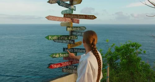 Woman traveler points to Kuala Lumpur on wooden directional signpost at Nusa Penida Bali coastal loo