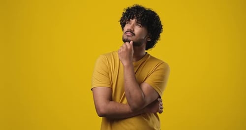 Pensive Young Indian Man Thinking with Hand Near Chin Standing Over Orange Studio Background