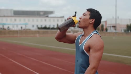 A Young and Fit Asian Guy Drinks Water From a Bottle After a Hard Workout at the Stadium Closeup