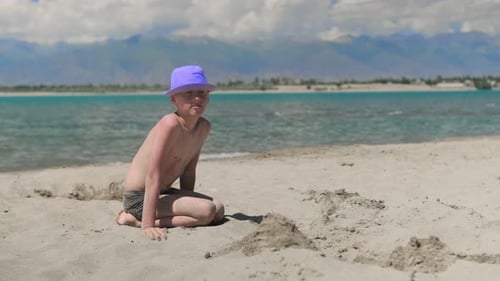 Little Boy in Purple Hat Playing with Sand on Beach Sea Lake with Mountains on Background Family