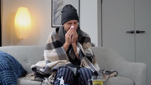 Un homme malade de la Reine des neiges est assis sur un canapé et porte un chapeau qui tient des mouchoirs et qui souffle un nez qui coule.