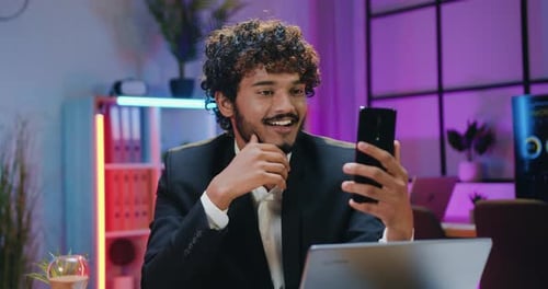 Handsome businessman enjoying smartphone video chat while overtime working in evening office.