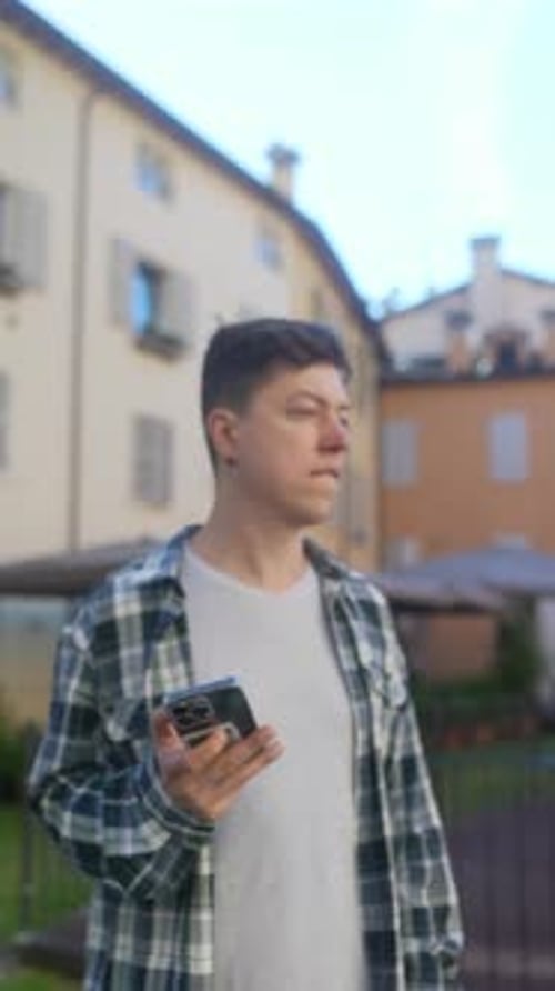 A Young Man in Urban Setting Holding Smartphone Looking Pensive Buildings in Background