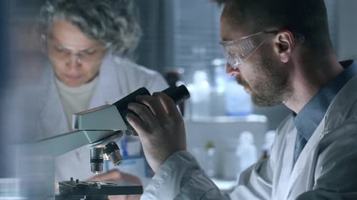 Wissenschaftler benutzt Mikroskop und macht sich Notizen, Kollegin arbeitet neben ihm