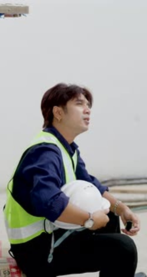 Asian male construction worker sits holding helmet inside unfinished building, resting after site