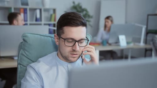 Handsome Office Worker in Black Eyeglasses Using Computer Talking on Mobile Phone While Working in