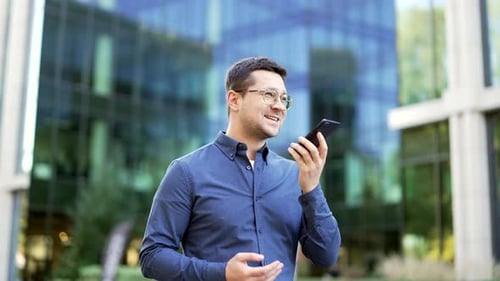 Businessman recording audio voice message on smartphone, holding phone near mouth walking on street