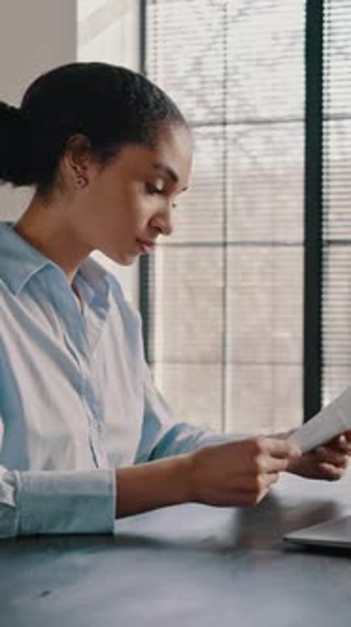 Purposeful Multiethnic Woman Carefully Reads Paper Contract Sitting at Desk in Office Interior