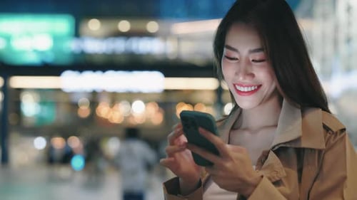 Positive Woman Holding Phone While Standing on the Street at Night and Scrolling Social Media Feed
