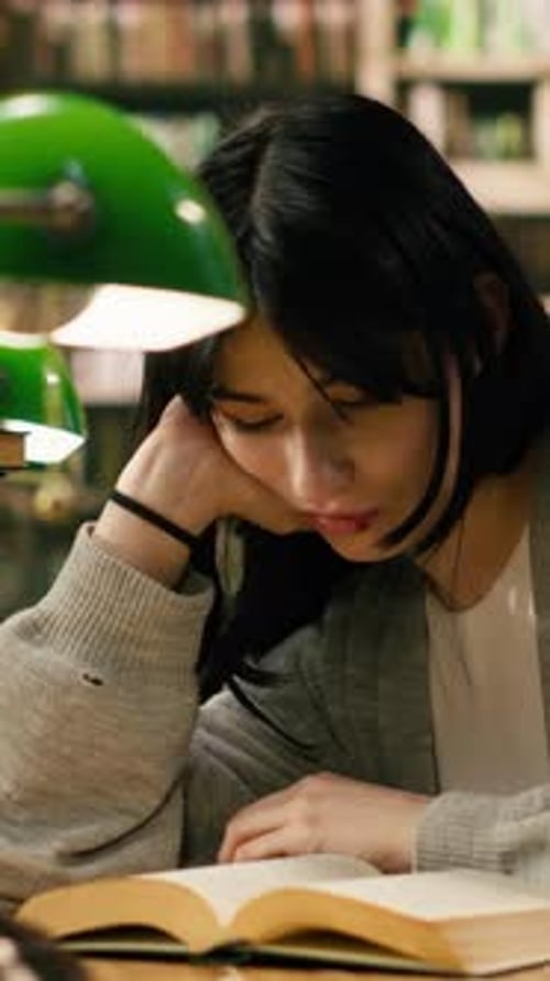 Vertical Video Tired Woman Student Leans on Her Hand While Reading Open Book Under Green Lamp