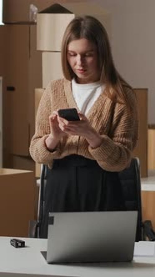 Young Woman Using Smartphone in Modern Home Office Workspace with Laptop and Cardboard Boxes