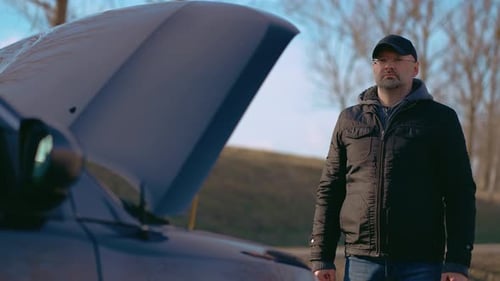 Confused Man Standing Near Broken Car with Open Hood and Damaged Engine Needs Help of Fix Services