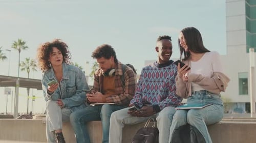 Students sit next to the university looking at their mobile phones and talking