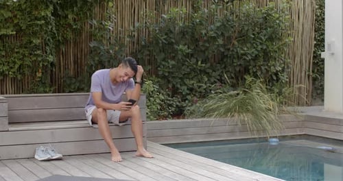 Sitting by pool, man smiling and using smartphone, relaxing outdoors, copy space
