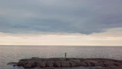 Man celebrates with elation standing on rocks looking out to empty sea horizon. Slow zoom in.