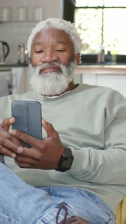 Vertical video: Holding phone, senior man reacting to message and laughing on couch at home
