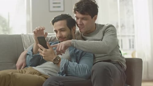 Cute Male Gay Couple Spend Time at Home. They are Lying Down on a Sofa and Use a Smartphone. They B