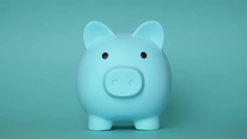 Hand inserting coin into light blue piggy bank on turquoise background