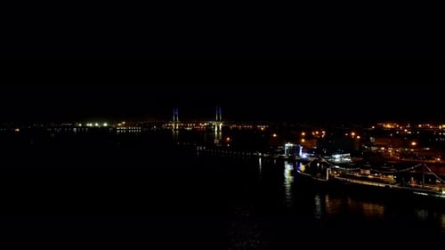 Night view of a bridge illuminated in blue with city lights and moored boats, reflected on water