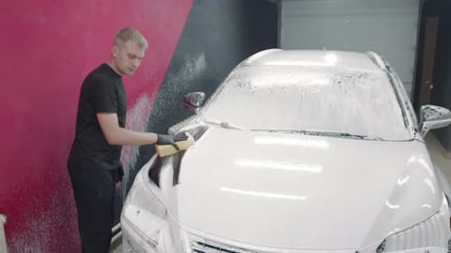 Caucasian Man Washes a Hood of a Sedan Auto Covered in White Foam with a Yellow Sponge