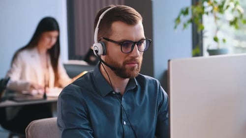 Ein Mann mit Kopfhörern mit Mikrofon arbeitet an einem Computer im Büro. Frau ist hinten