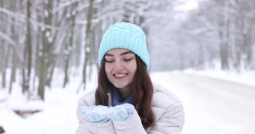 Beautiful woman blowing snow from hands in winter forest