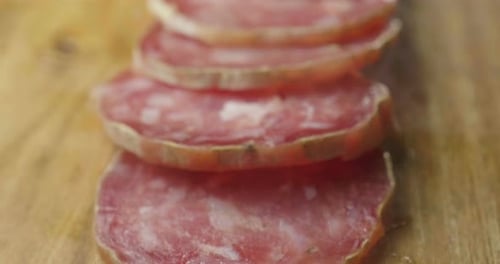 Macro shot of a salami on a wooden cutting board. One hand takes the knife and slices of slices to