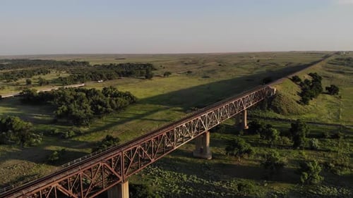 Sideways flyover above old rusty steel railroad track bridge in countryside at sunset, aerial