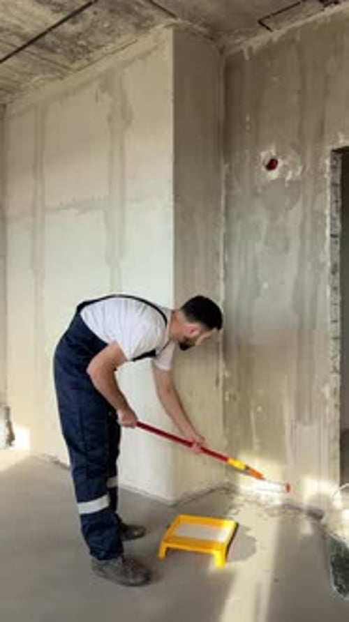 A Bearded Man in a Work Uniform is Priming the Walls After Applying Gypsum Plaster
