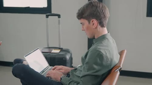 Jeune bel homme avec une valise assis travaillant son ordinateur portable dans le hall du terminal de l'aéroport