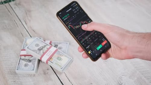 Hand Trades Stocks on Smartphone Near Bundles of Cash